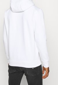 Sudadera blanca con capucha de textura suave y puños acanalados; combinada con unos jeans gris oscuro que presentan un pequeño logo en la parte trasera.