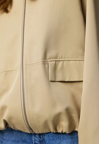 Beige jas met ritssluiting aan de voorkant, klepzak aan de rechterkant en elastische zoom, gedragen over blauwe spijkerbroek.