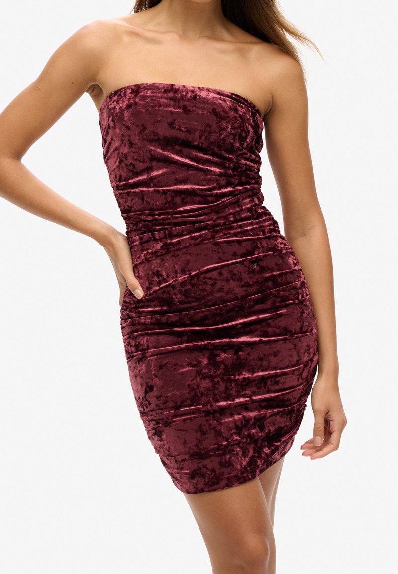 Femme portant une mini-robe bustier en velours rouge foncé froncée, une main sur la hanche, sur un fond clair uni.