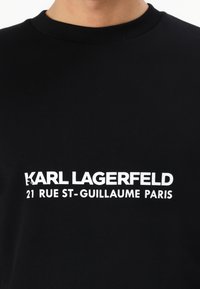 Crni pamučni dukserica s okruglim izrezom, koja sadrži podebljani bijeli tekst: "KARL LAGERFELD 21 RUE ST-GUILLAUME PARIS" istaknut.