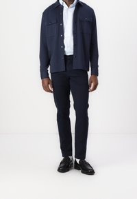 Marinblå skjortjacka med knappar och två framfickor, lager-på-lager över en ljusblå skjorta med krage, kombinerad med matchande byxor och svarta skor.