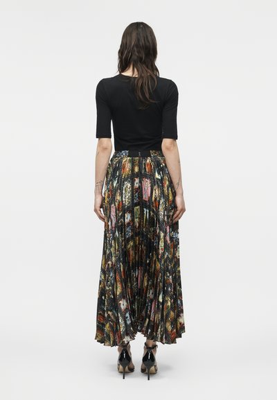 Kobieta o długich ciemnych włosach, ubrana w czarny krótki rękaw i kolorową, plisowaną, wzorzystą długą spódnicę maxi oraz czarne buty na wysokim obcasie.