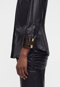 Vue du bras et de côté d'une personne portant une veste en cuir noire avec une large manchette en métal doré sur la manche et un pantalon en cuir froncé.