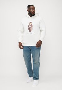 Sweatshirt branco com uma estampa de urso vestido de forma elegante, combinado com jeans azuis e sapatilhas brancas. O hoodie tem cordão e punhos canelados.