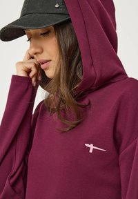 Bordeaux hoodie met een zachte textuur en lange mouwen, voorzien van een subtiele lichtroze logo-detail op de borst en een trekkoordkap.