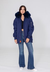 Navyblaue Steppjacke mit großem Pelzkragen, ausgestattet mit einem Reißverschluss vorne und Seitentaschen. Das Model trägt Schlagjeans und weiße Sneakers.
