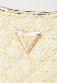 Keltainen tweed-laukku, jossa on kirjailtu pinta ja metalliset kultaiset yksityiskohdat. Sisältää kolmiomaisen kultaisen logoplaketin, jossa lukee "GUESS" ja kysymysmerkki.