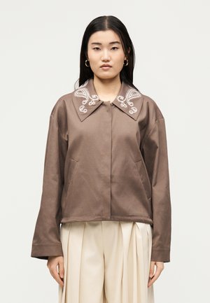 Mujer con chaqueta marrón con cuello bordado con diseño paisley y pantalones crema de pierna ancha y plisados, de pie frente a un fondo liso.