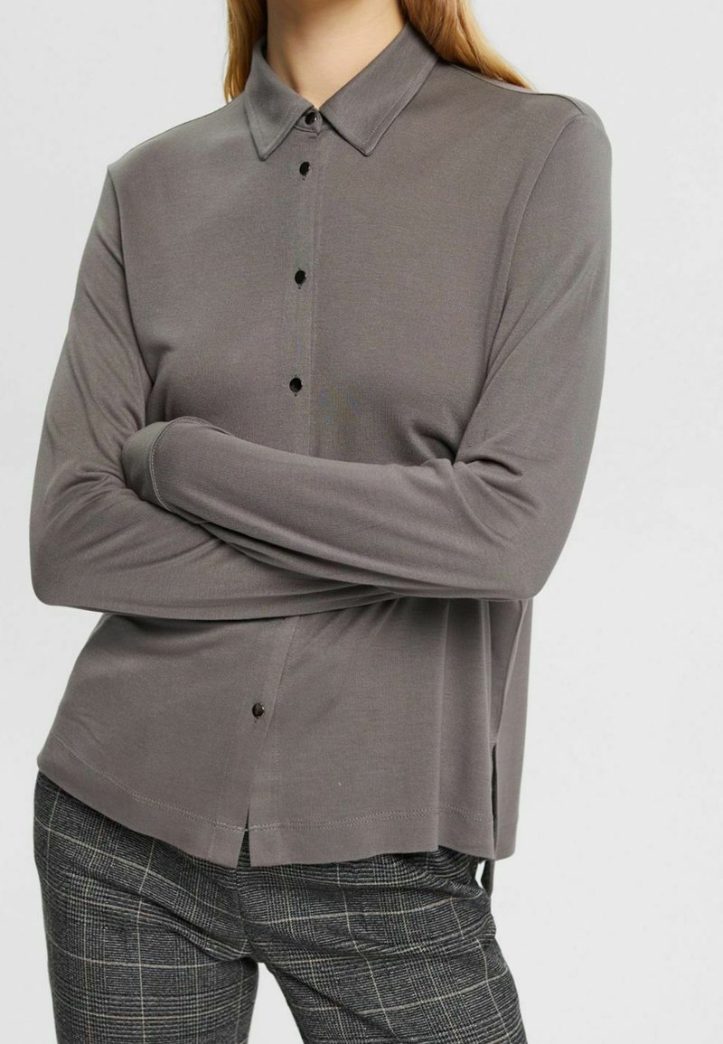 Chemise grise à col, à manches longues, en tissu doux avec un devant à boutons et des fentes sur les côtés. Portée avec un pantalon à motif tartan.