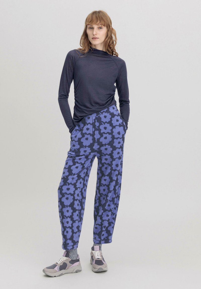 Langärmliges dunkles Oberteil mit gerafften Details, kombiniert mit lockeren, blumengemusterten lila Hosen. Die Schuhe haben graue und lilafarbene Akzente.