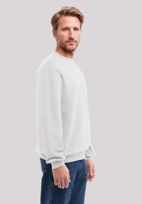 White cotton crewneck sweatshirt with ribbed cuffs and hem, paired with blue jeans. Model stands sideways, showing a relaxed fit and soft texture.