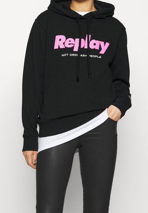 Person, die einen schwarzen Hoodie mit pinkem "Replay" Text und weißem "Not Ordinary People" Slogan, schwarze Hose und ein weißes Hemd darunter trägt.