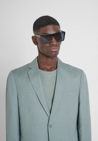 Blazer di lino grigio chiaro con rever a punta, chiusura a bottone singolo e una sottile tasca a toppa. Indossato sopra una t-shirt verde chiaro; occhiali da sole angolari scuri.