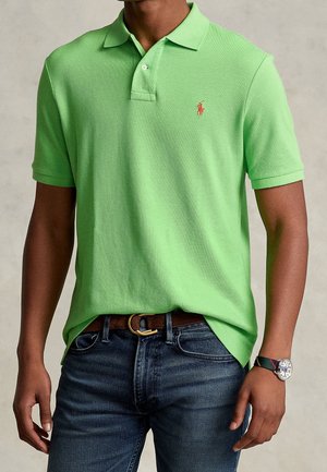 Mann mit hellgrünem kurzärmeligem Polohemd mit rotem Logo, dunklen Jeans, braunem Gürtel und einer Armbanduhr mit gestreiftem Armband.