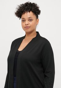 Μαύρο καναρίνι με v-neckline, μακριά μανίκια με διακοσμητικές λεπτομέρειες από στρας, απαλή υφή και ανοιχτό μπροστινό σχέδιο.