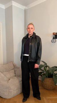 Crna kožna jakna preko tamne majice, crne široke hlače i crne cipele. Ima ovratnik, patentni zatvarač i minimalistički dizajn.