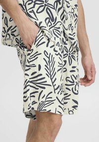 Hombre vestido con pantalones cortos y camiseta blanca con patrones abstractos de hojas y líneas en negro, con la mano en el bolsillo, de pie frente a un fondo liso.
