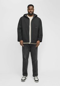 Veste à capuche noire avec fermeture éclair à l'avant, superposée sur une chemise beige clair. Portée avec un jean foncé et des baskets noir et blanc.