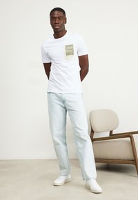 Hvit bomulls T-skjorte med grønn logo-trykk, paired med lyse denim jeans og hvite joggesko. En enkel, avslappet passform og uformelt design.