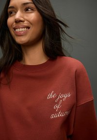 Bordový sveter s rebrovaným výstrihom a vyšitým textom "radosti jesene" v bielej farbe na hrudi. Hladká tkanina.