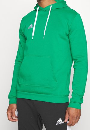 Mężczyzna w zielonej bluzie z kapturem Adidas z białymi sznurkami i czarnych spodniach Adidas, stojący na jednolitym tle.
