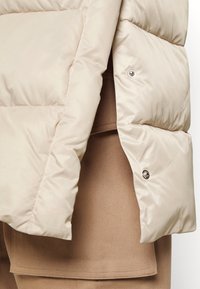 Veste matelassée beige avec boutons-pression métalliques ouverts sur un pantalon en tissu beige, montrant des détails et textures de vêtements superposés.