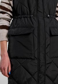 Svart kviltad väst med två framfickor och dragsnöre i midjan. Tillverkad av ett slätt, lätt material med matt yta.