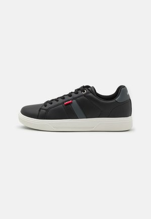 Zapatillas negras de material sintético con una suela de goma blanca. Presentan un diseño de cordones, detalles en gris y un logo de la marca en rojo.