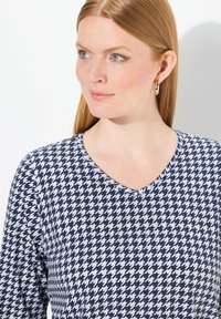 Oberteil mit Marine- und weißen Hahnentrittmuster, V-Ausschnitt und langen Ärmeln, aus einem weichen, glatten Stoff. Schlichte Details mit goldenen Creolen.