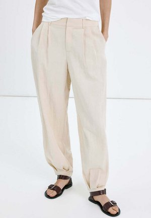 Persoon die losse beige broek met plooien draagt, handen in de zakken en donkerbruine open teensandalen met gespen tegen een effen achtergrond.