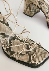 Hochhackige Sandalen mit einem schwarz-beigen Schlangenledermuster, ausgestattet mit kreuzweise angeordneten Riemen und einem quadratischen Zehenbereich. Strukturierte Oberfläche.