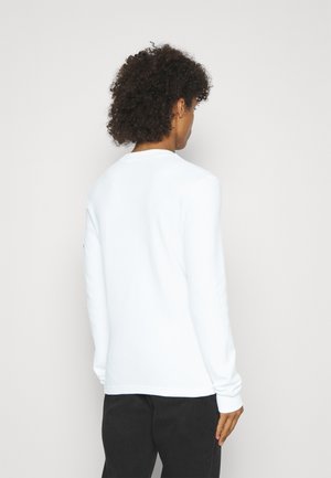 Mann med krøllete hår iført en enkel hvit langermet skjorte og svarte bukser, står vendt mot en lys grå vegg.