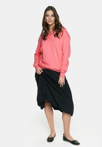 Różowy sweatshirt z długimi rękawami, zestawiony z czarną asymetryczną spódnicą. Model nosi czarne baleriny i srebrny pierścionek. Prosty, nowoczesny design.