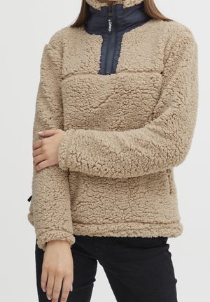 Beige, pluizige fleece trui met marineblauwe halve rits en opstaande kraag, gedragen met zwarte broek, armen over elkaar op de borst.