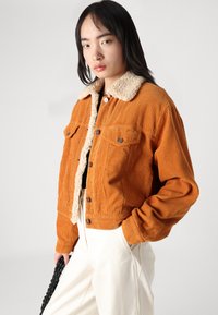 Veste en velours côtelé orange avec un col en fausse fourrure crème. Présente deux poches poitrine, une fermeture à boutons et un design court. Portée sur un pantalon blanc.