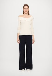 Top de malha canelada em off-white com design ombro a ombro, combinado com calças largas em azul-marinho e sapatos brancos sem atilhos. Look simples e moderno.