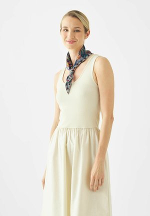 Vrouw met een mouwloze crèmewitte jurk en een sjaal met patroon om haar nek gestrikt, staand tegen een witte achtergrond.