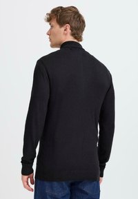 Pull noir à manches longues avec col montant, poignets côtelés et ourlet. Le tissu a l'air lisse avec une texture subtile, porté sur un jean bleu.