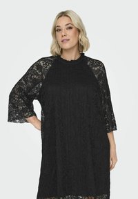 Czarna koronkowa sukienka z falbaniastym dekoltem, trzema czwartego rękawami i luźnym krojem. Koronka ma kwiatowy wzór i teksturowane wykończenie.