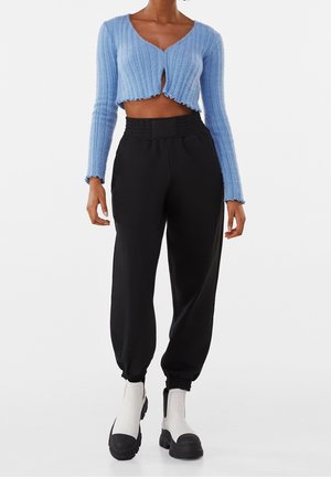 Jasnoniebieski krótki kardigan o prążkowanej fakturze z wycięciem w kształcie klucza, zestawiony z czarnymi, zwężanymi dresami i białymi botkami za kostkę.