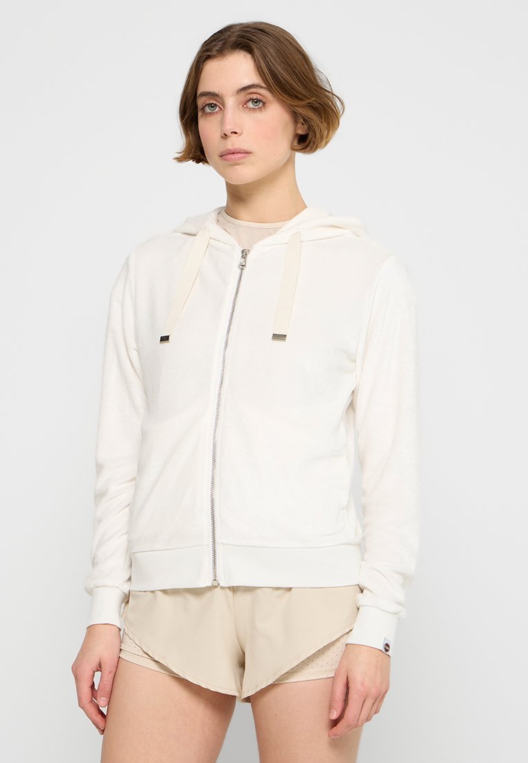 Felpa bianca con zip in morbido tessuto con cappuccio, dotata di cordini beige e hardware argentato, abbinata a pantaloni corti beige.