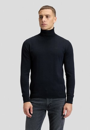 Uomo con capelli castani corti che indossa un maglione dolcevita nero aderente e jeans scuri, in piedi davanti a uno sfondo grigio chiaro e uniforme.