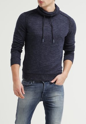 Man draagt een marineblauwe gebreide trui met aantrekkoord bij de kraag en vervaagde blauwe spijkerbroek, hand in zak, tegen een effen lichte achtergrond.