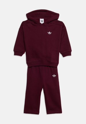 Sötétvörös, kisgyermek méretű kapucnis pulóver és nadrág szett, fehér Adidas trefoil logókkal a mellrészen és a nadrágszáron, fehér háttéren laposan elhelyezve.