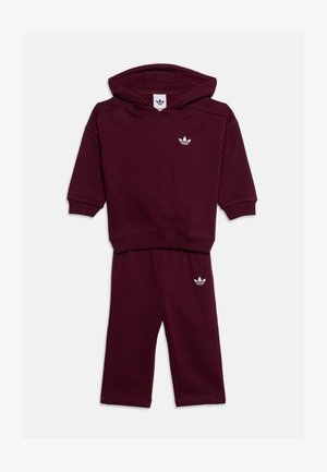 Sötétvörös, kisgyermek méretű kapucnis pulóver és nadrág szett, fehér Adidas trefoil logókkal a mellrészen és a nadrágszáron, fehér háttéren laposan elhelyezve.
