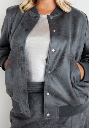 Femme portant une veste bomber grise texturée sur un top blanc côtelé, les mains dans les poches de la veste.