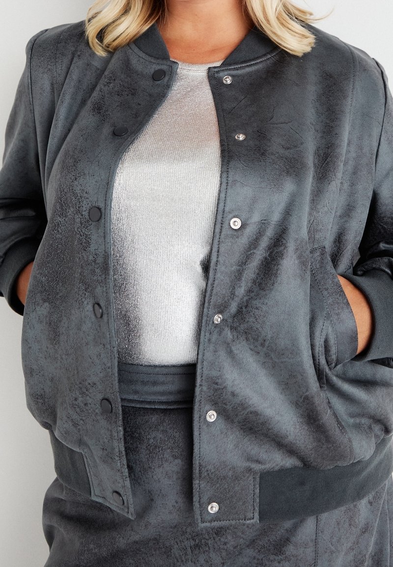 Femme portant une veste bomber grise texturée sur un top blanc côtelé, les mains dans les poches de la veste.