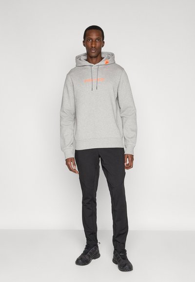Mann trägt hellgrauen Kapuzenpullover mit orangefarbenem Text, schwarze Hose und schwarze Sneaker, steht vor einem einfarbigen weißen Hintergrund.