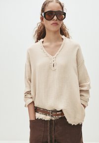 Beige, strukturiertes Langarmoberteil mit V-Ausschnitt und Rüschendetails, kombiniert mit braunen Hosen und einem genagelten Gürtel. Große schwarze Sonnenbrille.