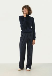 Marineblauer Rollkragenpullover mit gerippten Bündchen, kombiniert mit weit geschnittenen marineblauen Hosen und weißen Schuhen mit holzähnlichen Sohlen. Einfaches, schlichteres Design.